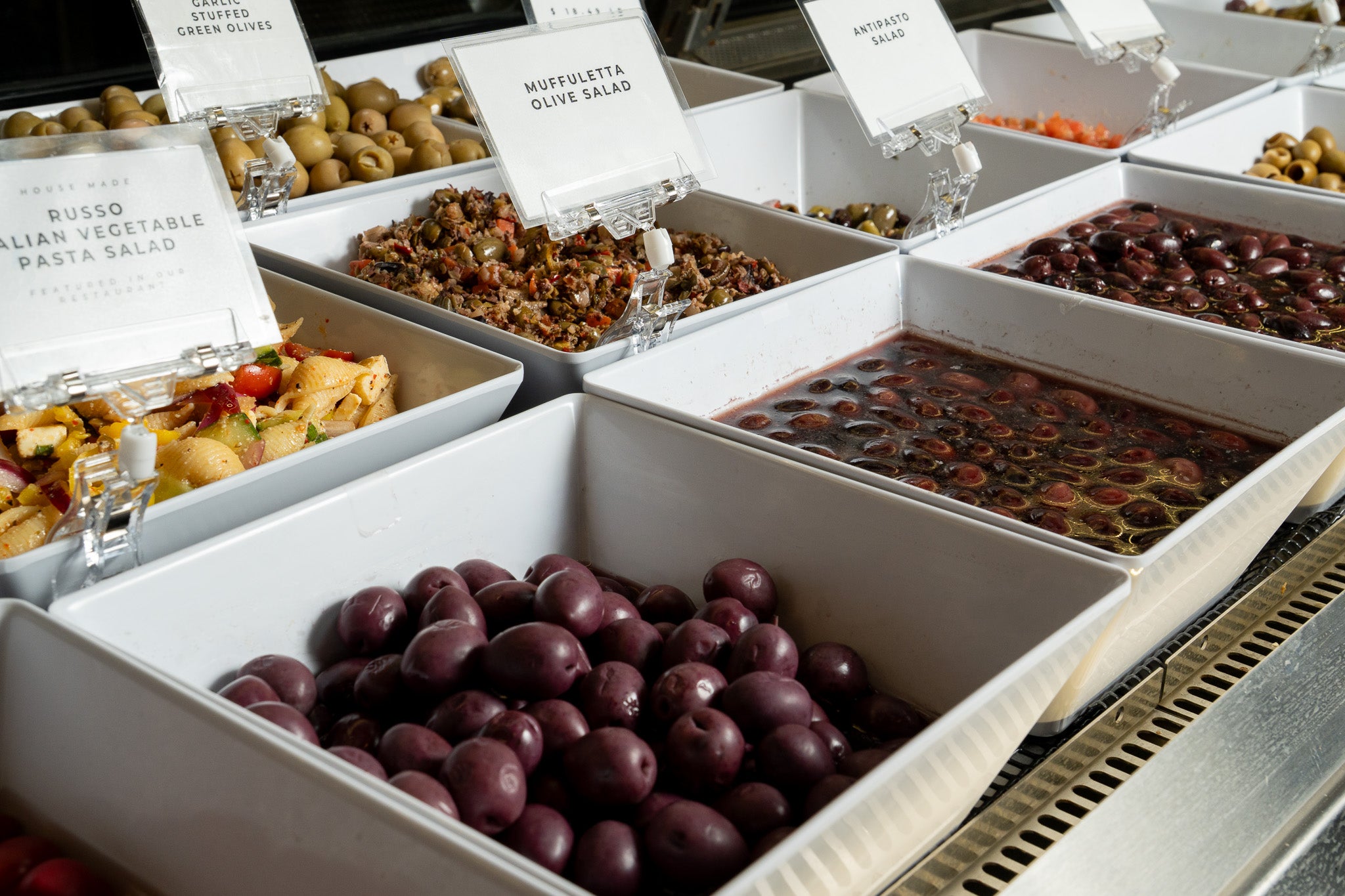 Assorted olives and other food items displayed in a deli case setting with labels.
